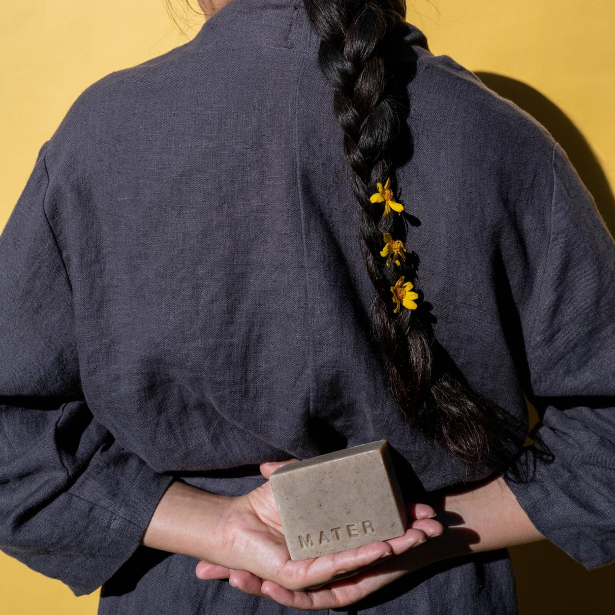 Woman with a long black hair braid and her back to the camera holding an unwrapped bar of Mater Soap.
