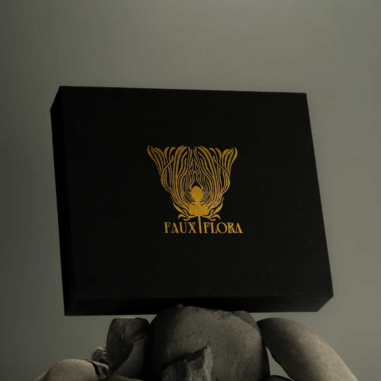 Black box with gold Faux Flora logo and text held by a rocks against a gray background