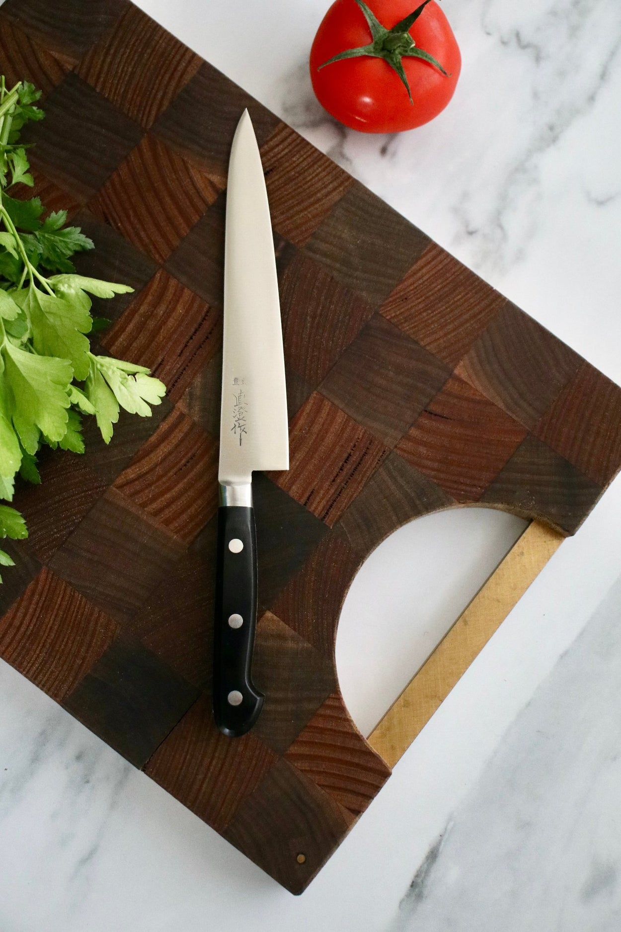 Japanese Naozumi Nihonkou 15cm Sharp Carbon Steel Kitchen Knife with tomato and parsley on wood cutting board, on marble bench.