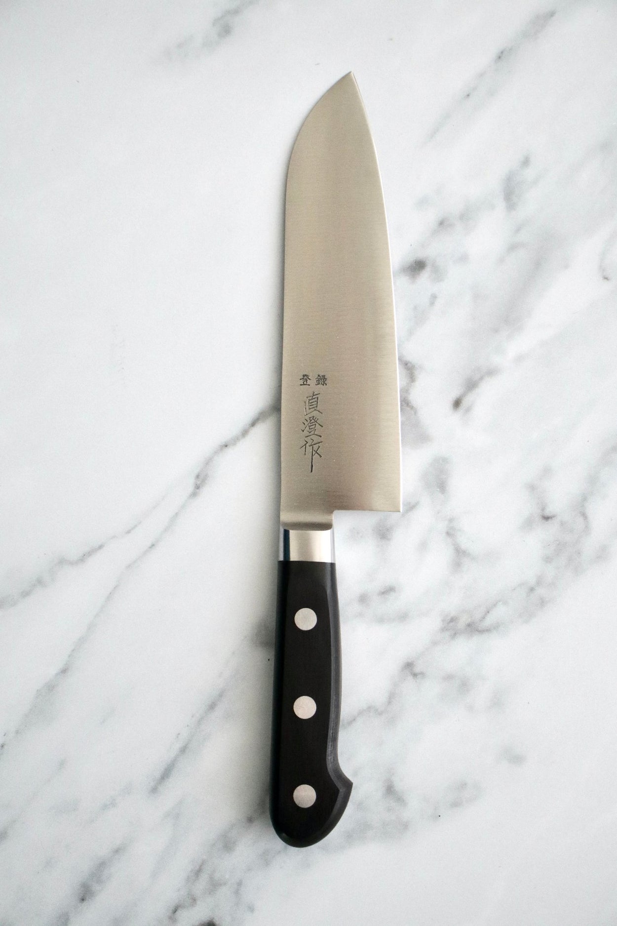 Japanese Naozumi Nihonkou 165mm Santoku Carbon Steel Kitchen Knife on chopping board with tomato, parsley and marble background.