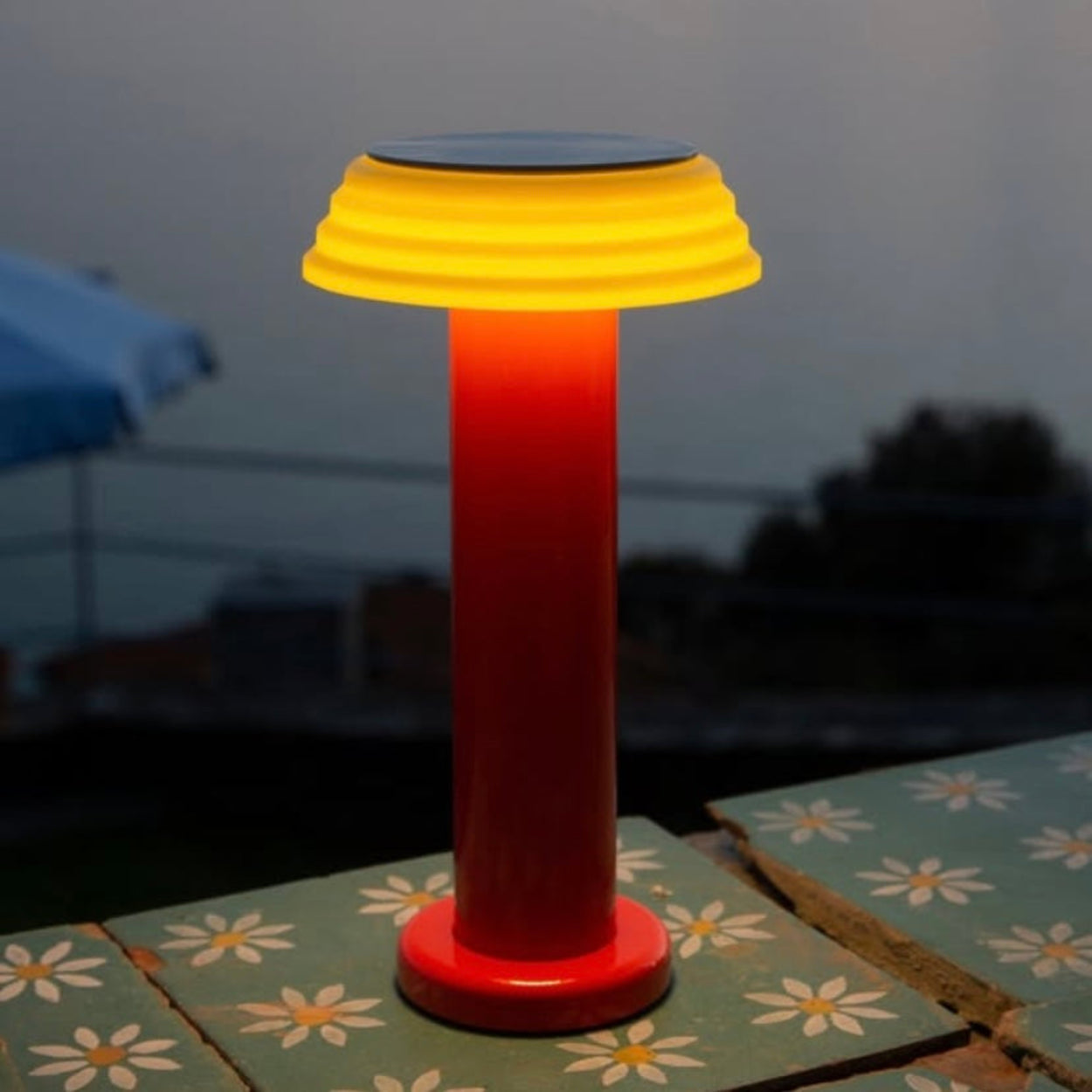 Sowden PL1 rechargeable outdoor lamp with yellow top and red base on a table with a floral pattern.