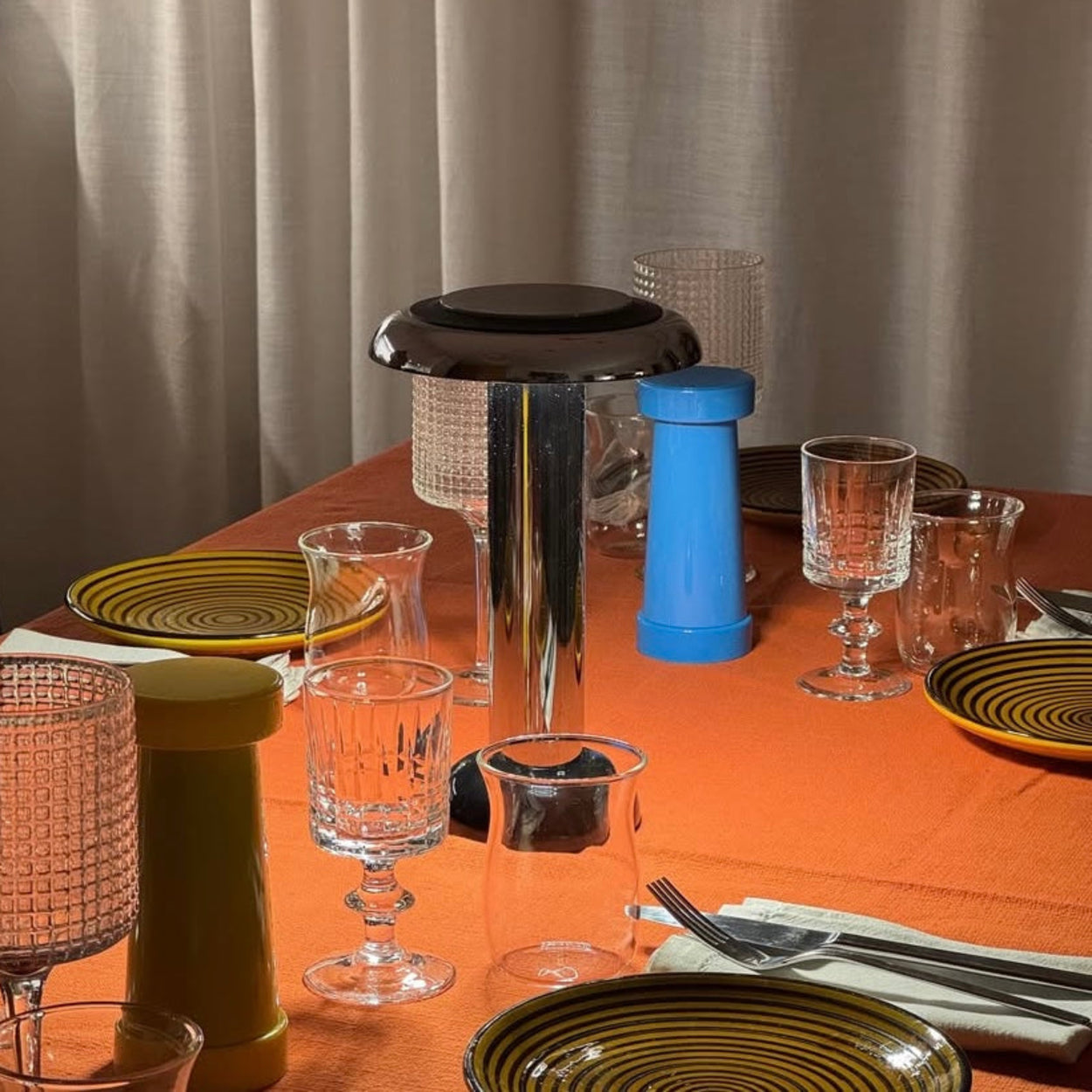 Dining table setting with Sowden Pl6 lamp, plates, glasses, and cutlery on an orange tablecloth.
