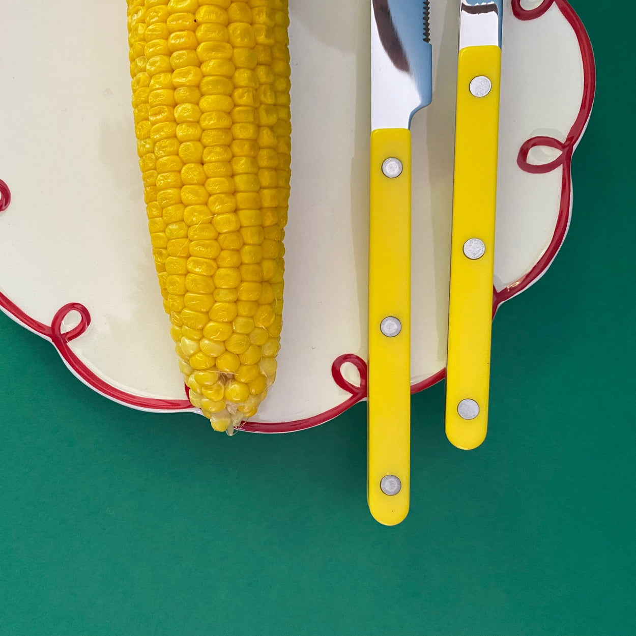 Sabre Paris Bistrot 4 piece cutlery set in yellow on a white and red squiggle plate, with corn cob, against a green background.