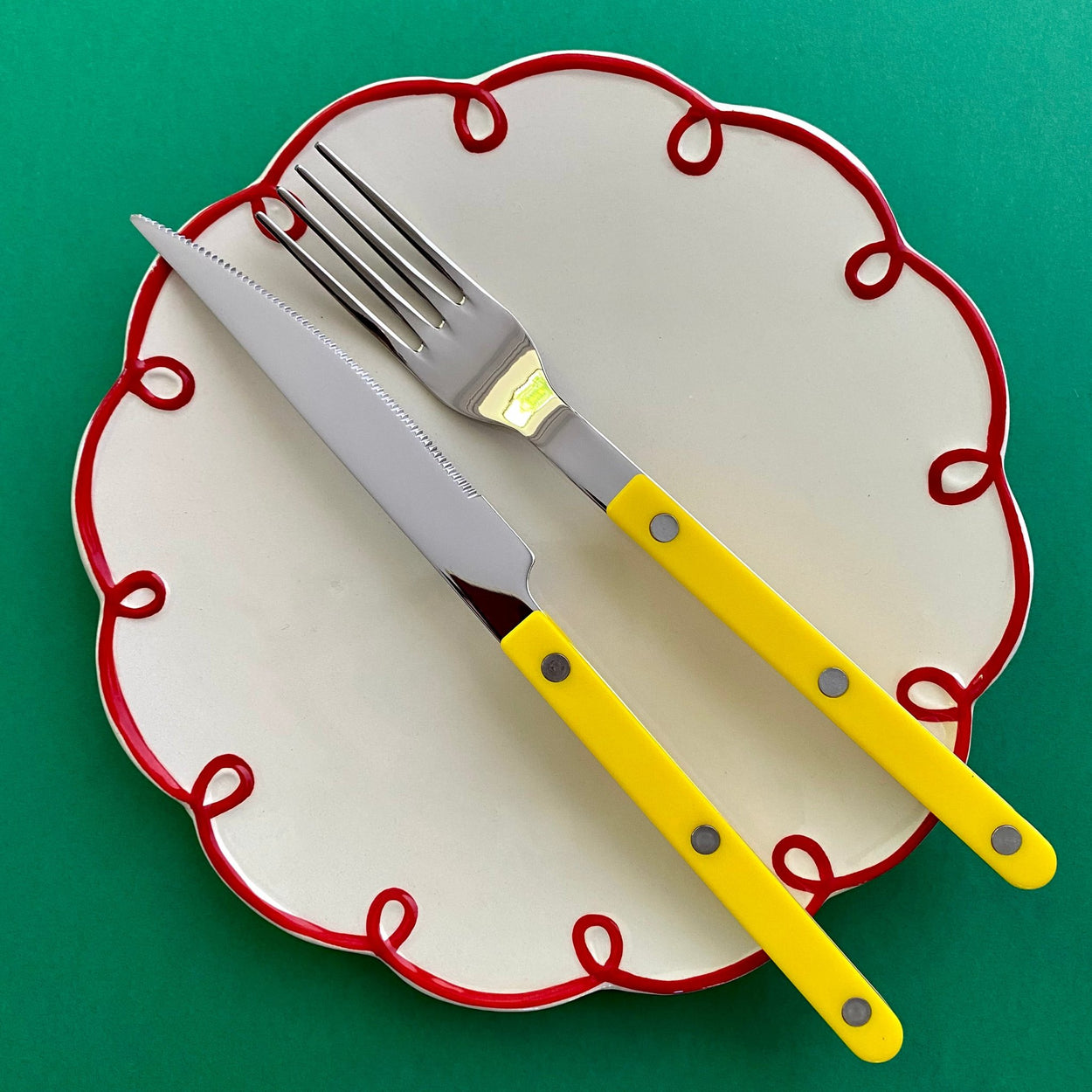 Sabre Paris Bistrot 4 piece cutlery set in yellow on a white and red squiggle plate against a green background.