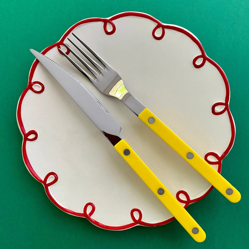 Sabre Paris Bistrot 4 piece cutlery set in yellow on a white and red squiggle plate against a green background.