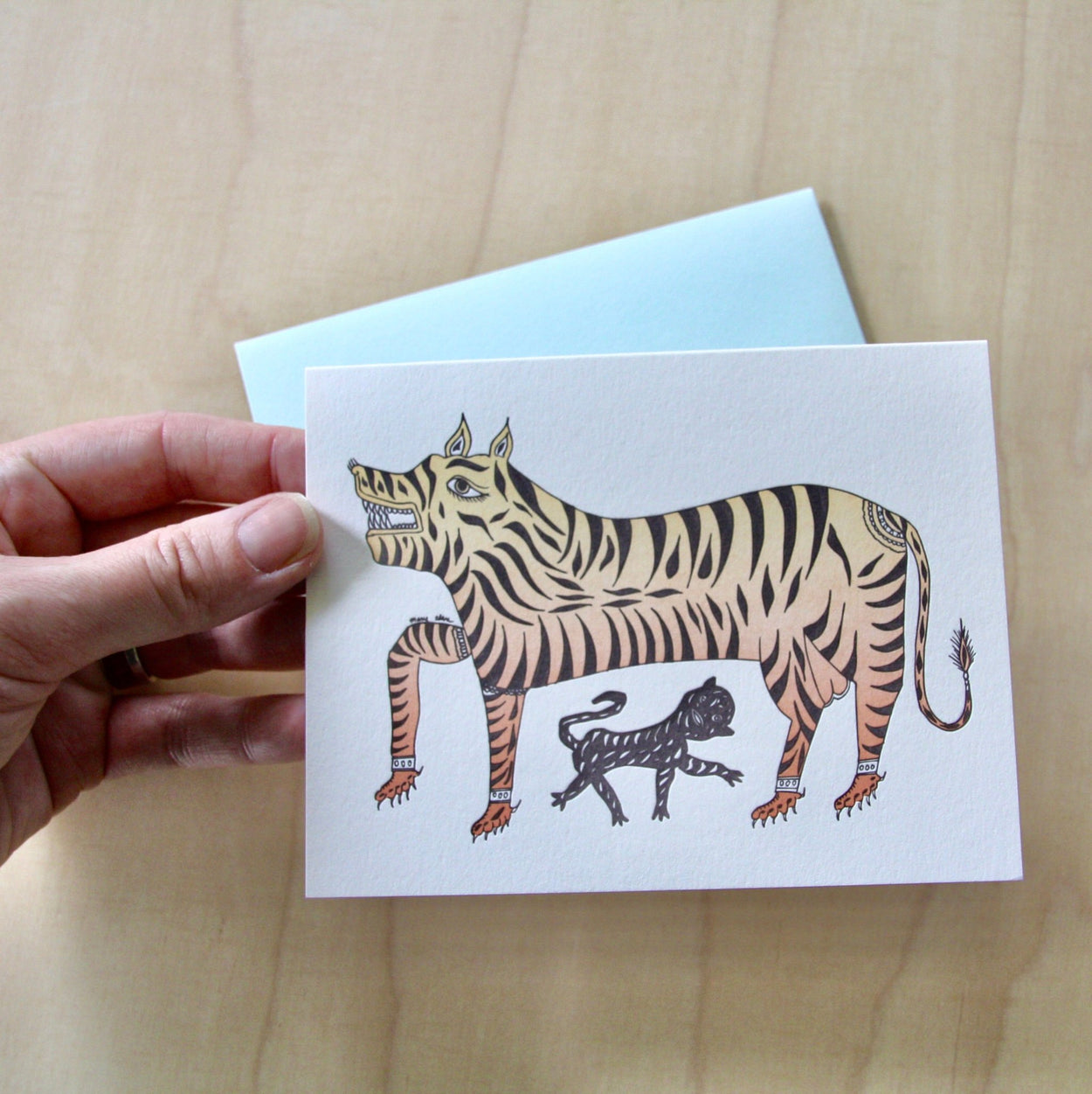 Hand holding Tiger Letterpress Greeting Card with pale blue envelope on pale wood.