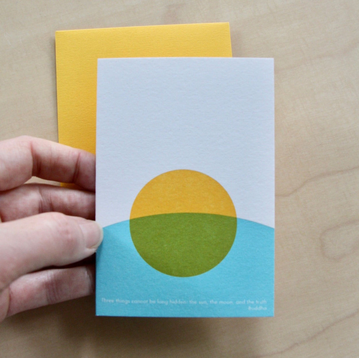 Hand holding a Hand printed greeting card with yellow sun and blue curve on a yellow envelope. reading "Three things cannot be long hidden, the sun, the moon and the truth" Buddha