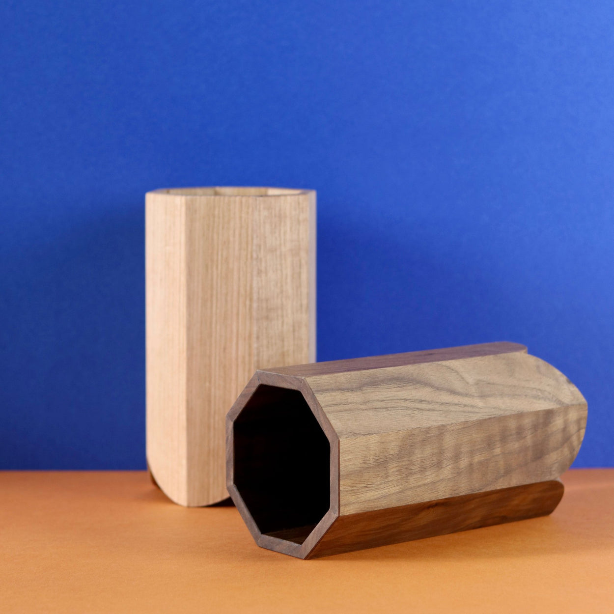 Handmade Wood Kitchen Utensil Holder in walnut lying on its side in front of the Oak Handmade Kitchen Utensil Holder, against a blue and brown background.