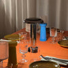 Sowden Lamp PL6 in Chrome on a orange dining table with glassware plates and cutlery with beige curtain backdrop.