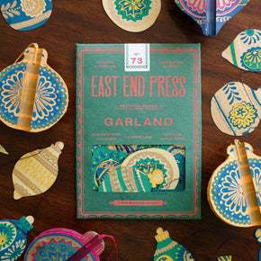 Garland by East End Press surrounded by decorative paper ornaments on a wooden surface.
