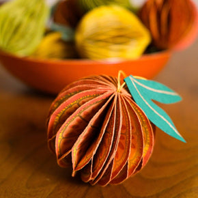 Decorative paper orange with leaf on a wooden surface, surrounded by colourful pumpkins in the background.