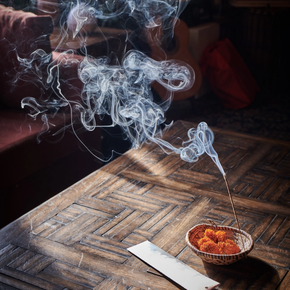 A small woven bowl filled with orange incense sticks sits on a dark wooden table, with smoke curling upward in a dimly lit interior setting.