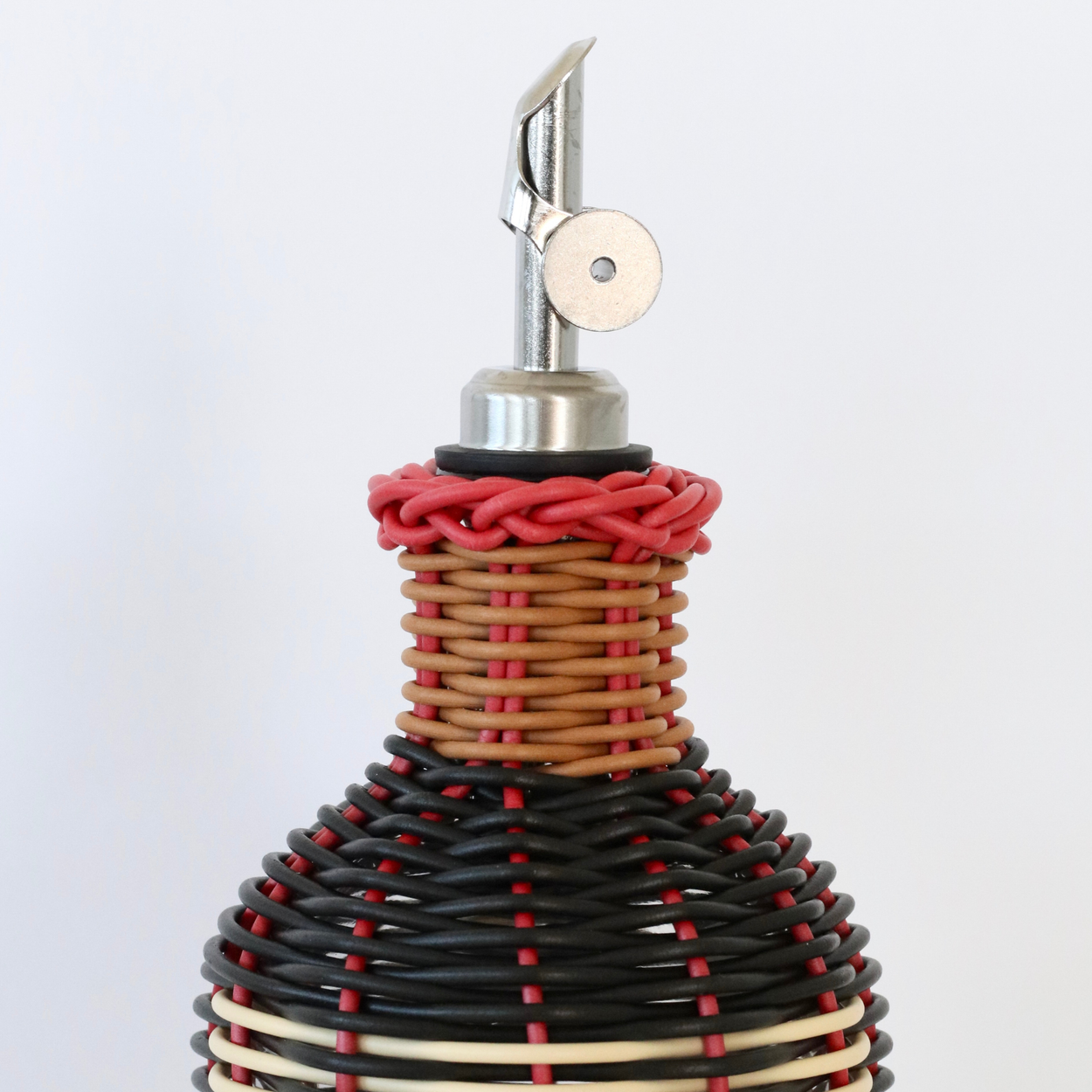 Close up of nozzle of decorative olive oil bottle with black, red and cream woven design and metallic nozzle on a white background