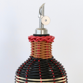 Close up of nozzle of decorative olive oil bottle with black, red and cream woven design and metallic nozzle on a white background