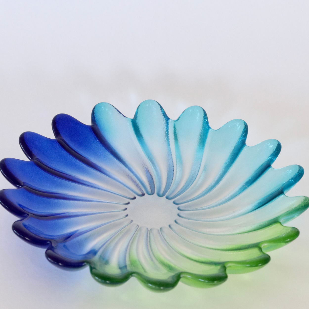 A scalloped glass bowl with a petal-like design, featuring a blue and green gradient transitioning through light blue in the center, set against a plain light background.