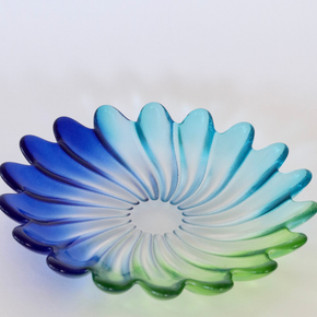 A scalloped glass bowl with a petal-like design, featuring a blue and green gradient transitioning through light blue in the center, set against a plain light background.