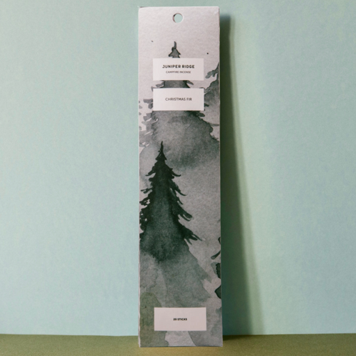 A tall rectangular incense box standing upright against a pale blue wall. The front features a watercolor-style forest scene in gray, black, and white, with small white label areas containing text including 'Juniper Ridge Christmas Fir'