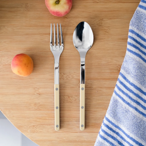 A stainless steel Sabre Paris Serving set with cream-coloured handles, featuring a fork and a spoon, arranged on a light wood surface next to a blue and white striped cloth and two peaches.