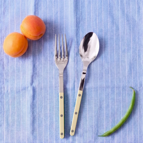 A flat-lay view of a silver metal Serving Set fork and spoon with pale white handles, set on a blue and white striped cloth draped over a wooden chair. Two apricots and a green bean are placed nearby on the cloth, with a green chalkboard background.