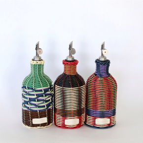 Three woven bottle-shaped oil or vinegar dispensers with metal pour spouts, shown side by side on a plain light background. Each dispenser has a different multicolor wrap pattern: green, black and red, and red, blue, and purple.