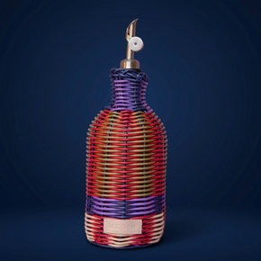 A woven bottle cover or decorative bottle wrap in a multicolour wicker pattern with purple, red, orange, green, and cream stripes, featuring a silver-toned pour spout and a small beige brand patch that reads 'MAEGEN'.