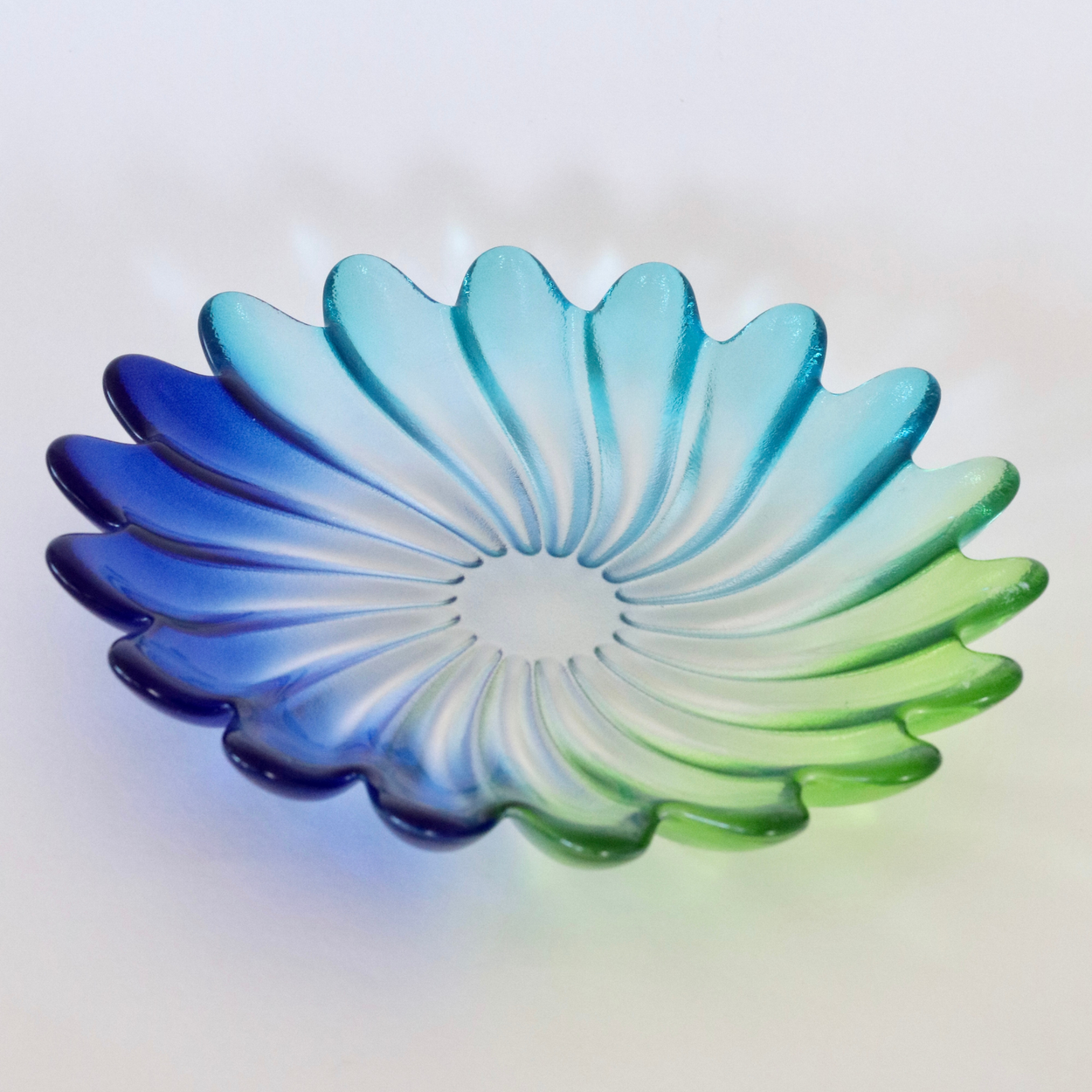A scalloped glass dish with a petal-like design, featuring a gradient color transition from deep blue on the left to light cyan in the center and bright green on the right, set against a plain white background.