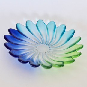 A scalloped glass dish with a petal-like design, featuring a gradient color transition from deep blue on the left to light cyan in the center and bright green on the right, set against a plain white background.
