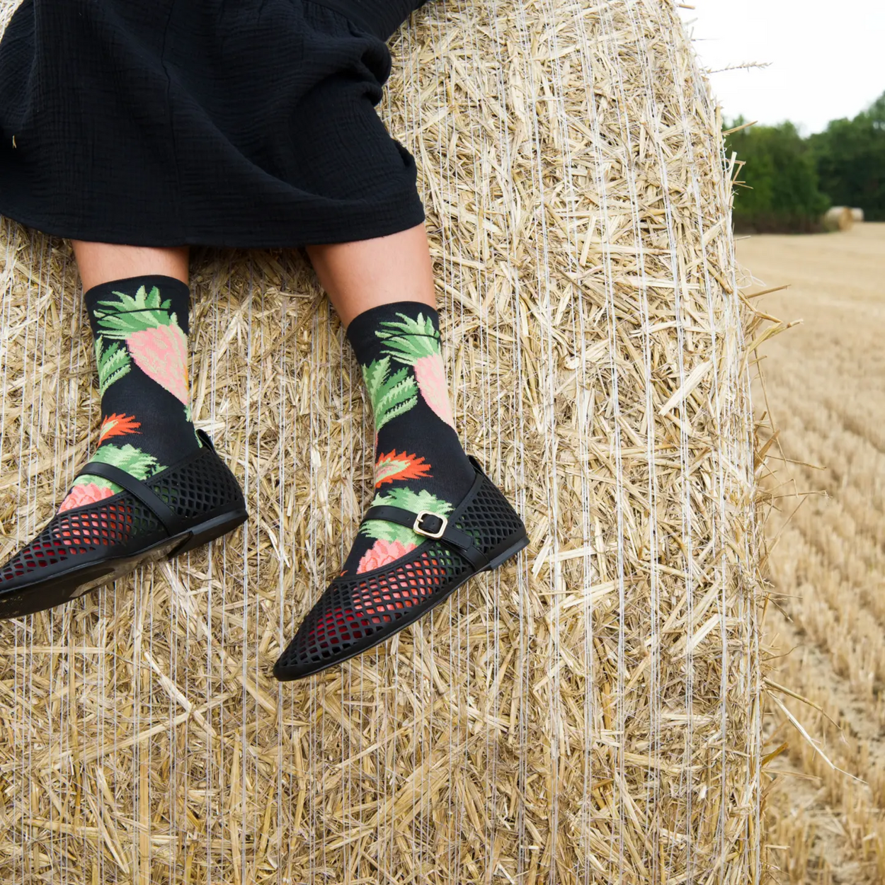 Woman on hay bale with black skirt and shoes and Bonne Maison pineapple socks.