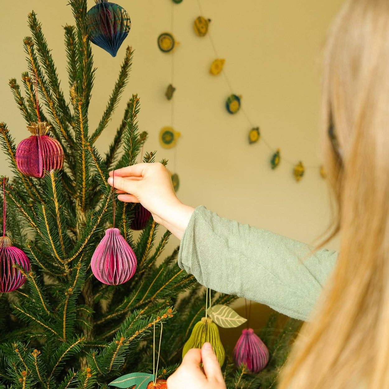 Person decorating a Christmas tree with colorful ornaments in a cozy room.