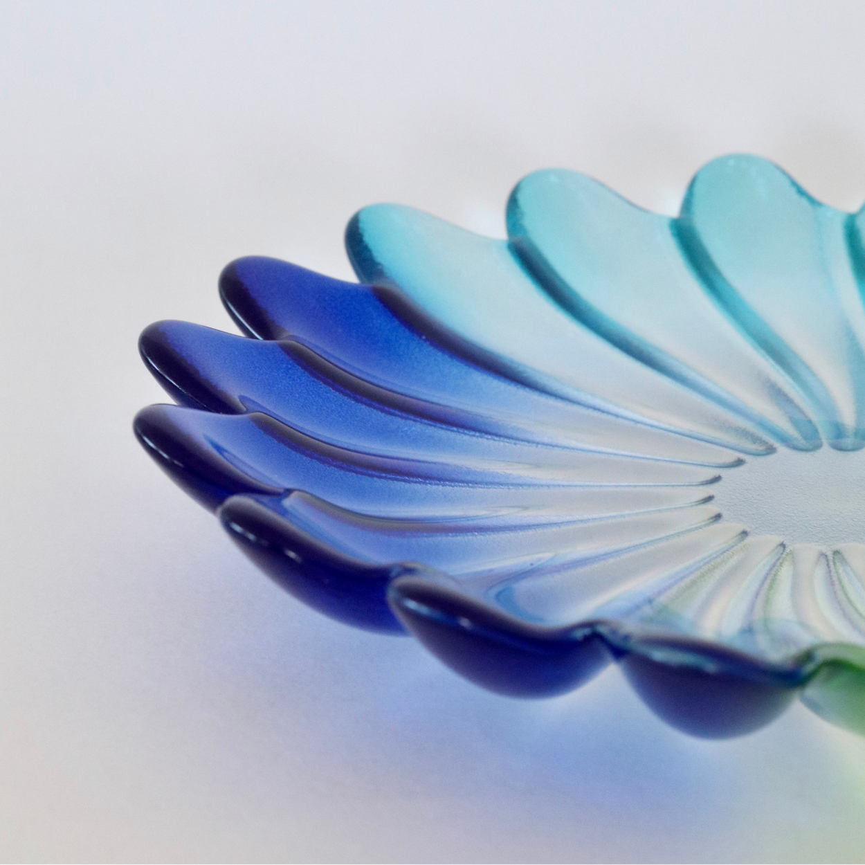 A close-up of a translucent scalloped glass dish or plate in a blue-to-turquoise gradient, photographed on a clean white background.