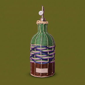 A decorative bottle with a woven wicker-style body in green, purple, brown, and beige, topped with a metal pour spout and a small white circular detail, shown on an olive green background.