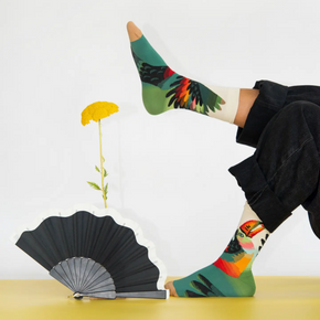 Person in black pants wearing Bonne Maison Toucan socks on white backdrop with fan and yellow flower