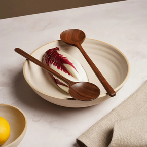 2 walnut handmade serving spoons resting on a beige ceramic bowl with lettuce and beige linen napkin. all on a marble table top.