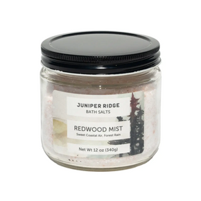 A 340g glass jar of Juniper Ridge Bath Salts in the Redwood Mist scent, featuring pinkish salt crystals inside and a black screw-on lid, shown on a plain white background.