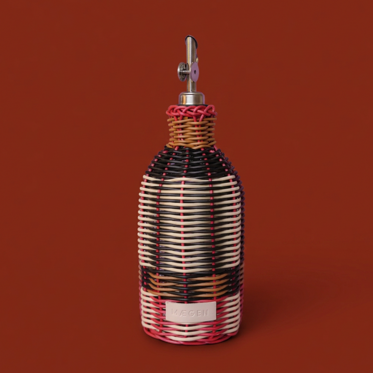 A decorative bottle with a woven wicker-style body in black, white, and red, topped with a silver metal pour spout, set against a solid rust-brown background.