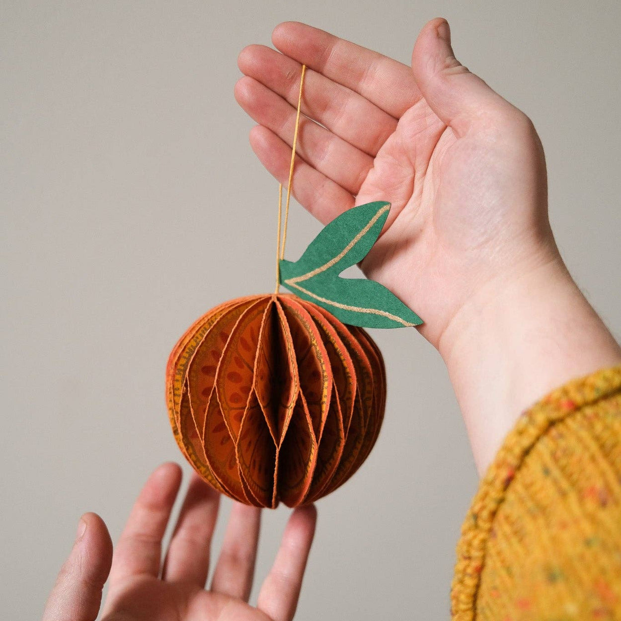 Hand holding a decorative paper fruit ornament against a neutral background