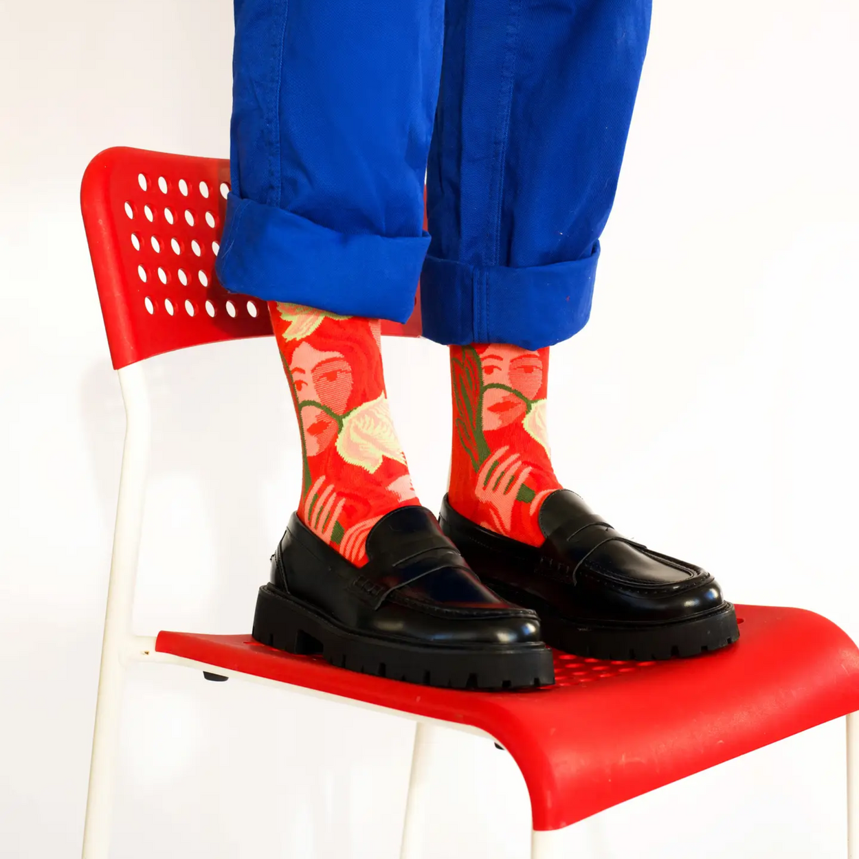 Bonne Maison Red Visage socks worn by person with blue pants standing on a red chair.