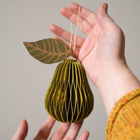 Hand holding a decorative paper pear ornament with a leaf against a neutral background