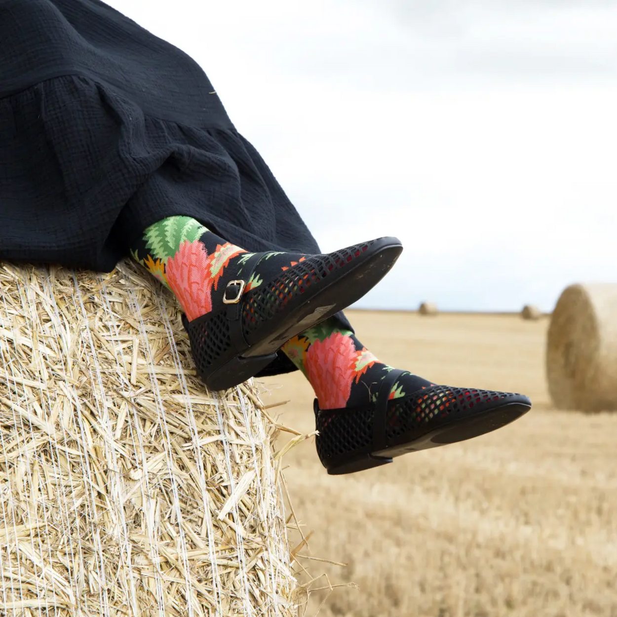 Woman on hay bale with black skirt and shoes and Bonne Maison pineapple socks.
