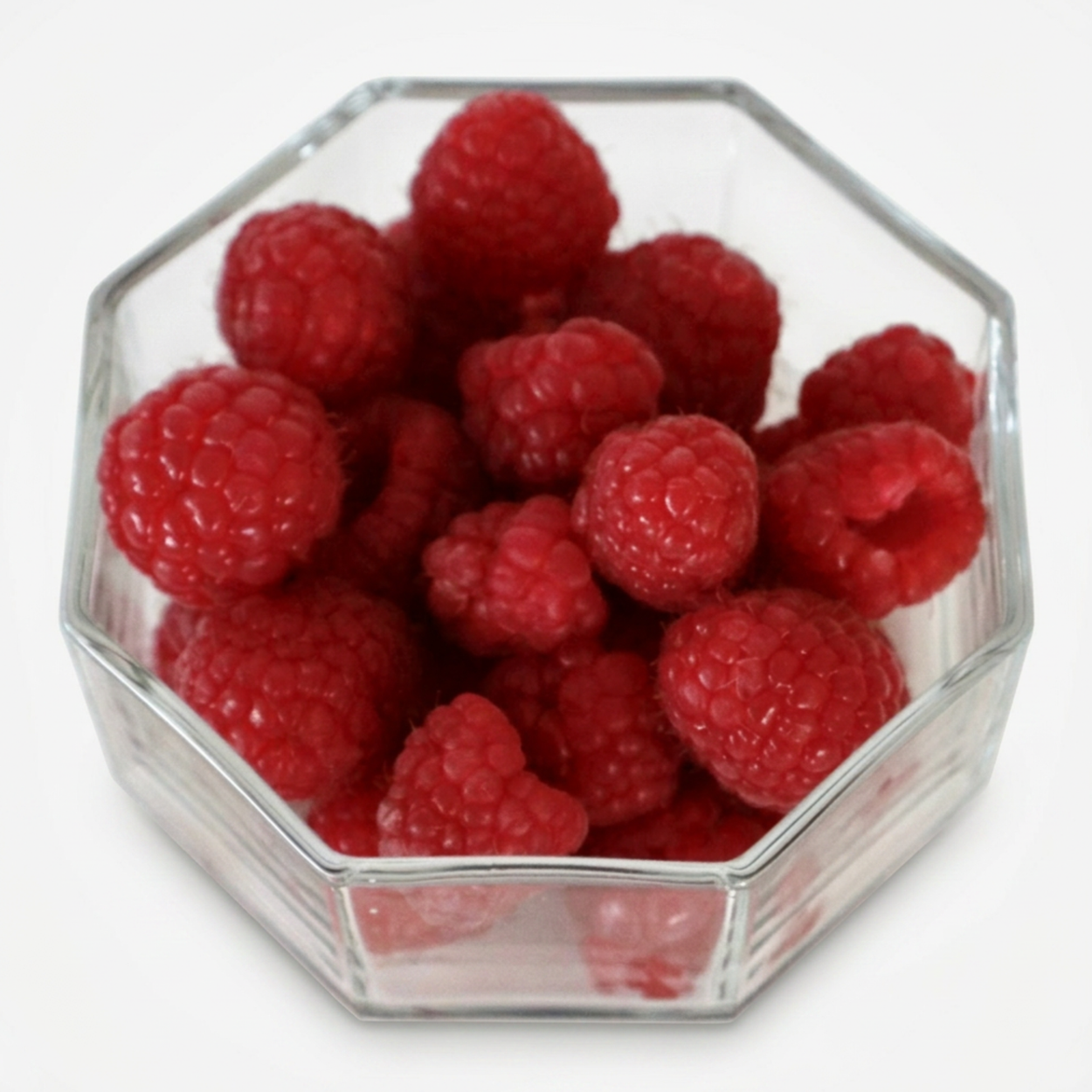 Hexagonal glass bowl filled with red raspberries on a white background