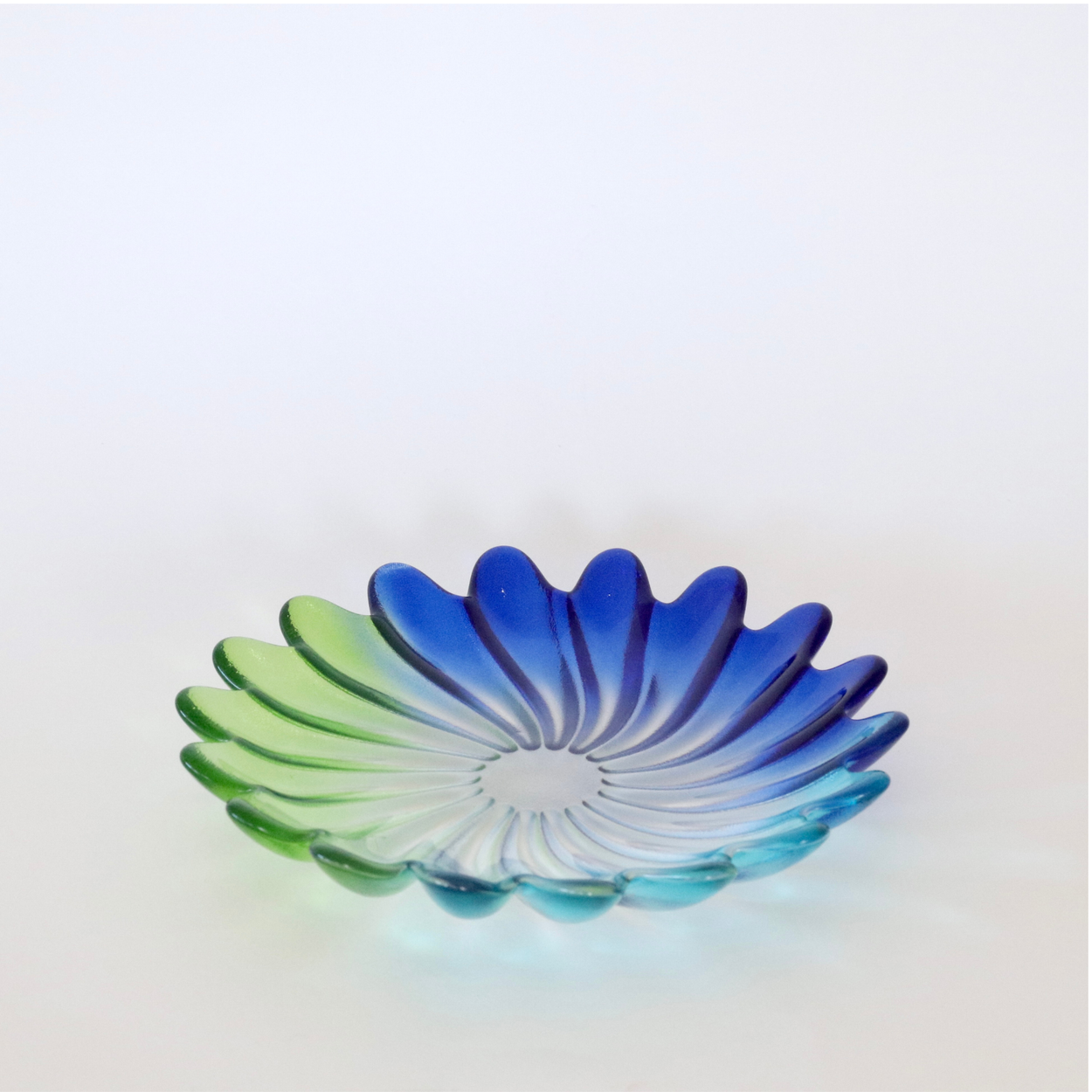 A shallow petal-shaped glass bowl with a blue-to-green gradient, photographed on a clean white background.