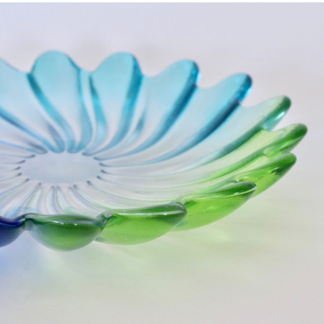A translucent glass dish with a petal or flower-like scalloped rim, shown in a close-up studio shot. The glass has a color gradient transitioning from light blue at the top to green at the bottom, set against a plain white background.