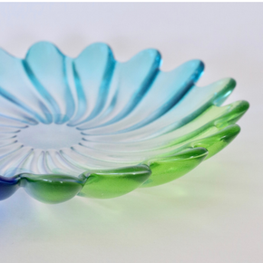 A translucent glass dish with a petal or flower-like scalloped rim, shown in a close-up studio shot. The glass has a color gradient transitioning from light blue at the top to green at the bottom, set against a plain white background.