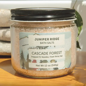 A glass jar of Juniper Ridge Cascade Forest bath salts with a black screw-on lid, sitting on a wooden surface with a blurred background.