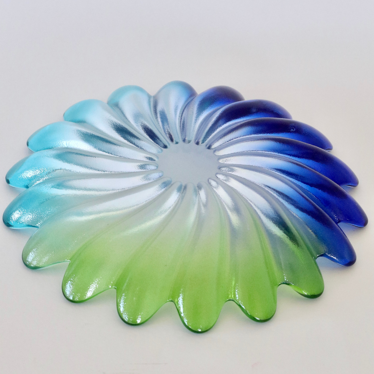 A circular, petal-shaped glass decorative piece with a gradient color transition from light blue and green to deep blue, shown on a plain light background.