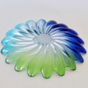 A circular, petal-shaped glass decorative piece with a gradient color transition from light blue and green to deep blue, shown on a plain light background.