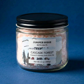 A glass jar of Juniper Ridge Bath Salts, 'Cascade Forest' scent, filled with pinkish salt crystals and sealed with a black lid, set against a dark blue background.