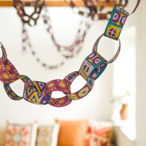 A colorful patterned paper chain hanging in a bright, modern living room with a blurred sofa and pillows in the background.