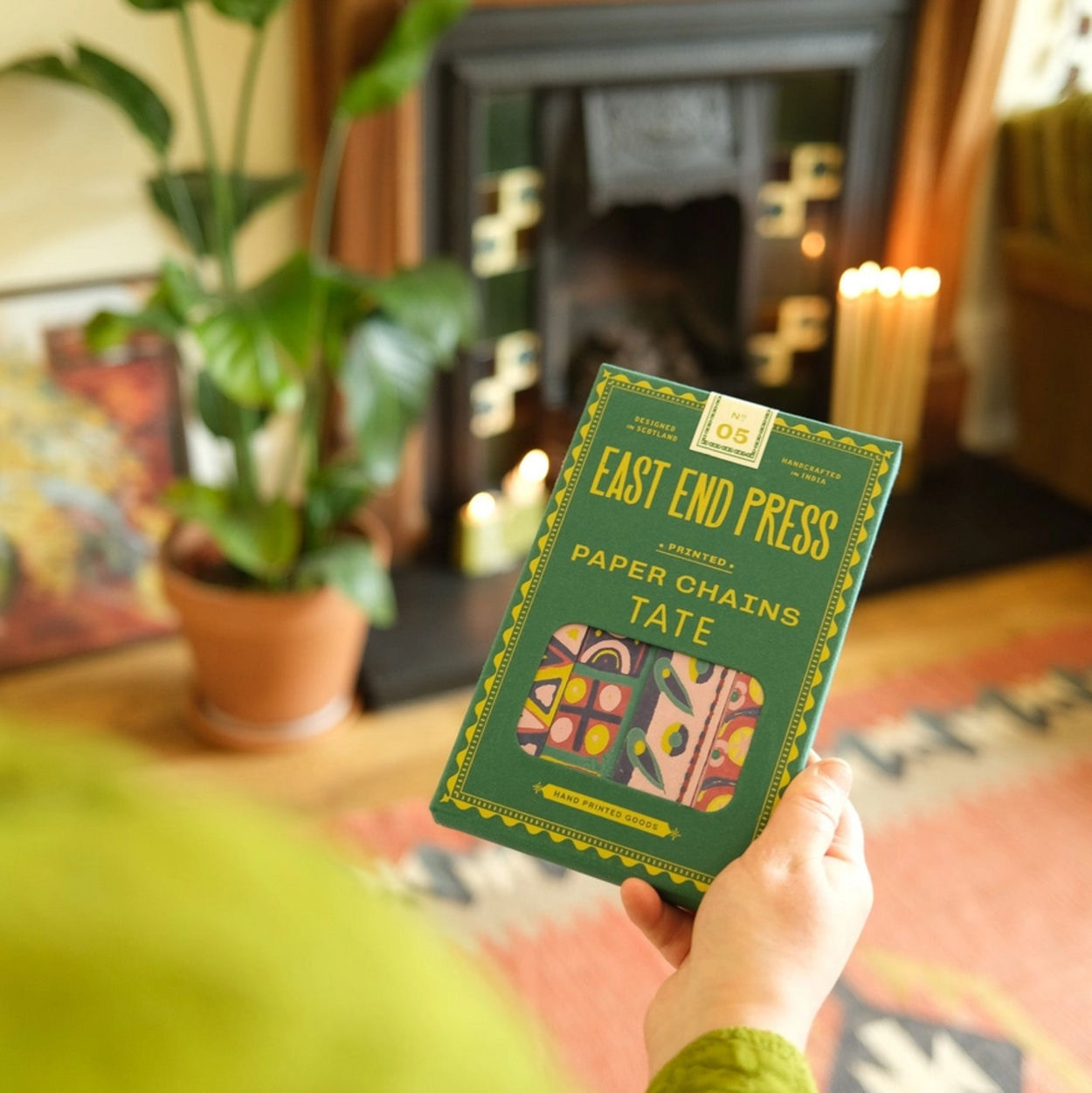 Person holding a Paper Chain kit by 'East End Press' in a cozy room with a fireplace and plants.