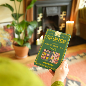 Person holding a Paper Chain kit by 'East End Press' in a cozy room with a fireplace and plants.